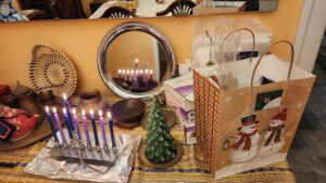 A lit menorah filled with blue and purple candles, reflected in a silver plate, next to a little model Christmas tree, next to a gift bag with snow people wearing scarves and hats